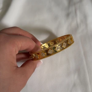  Van cleef Guldigt armband med blommönster - Snyggt armband i guldton med glittriga små stenar i blommönster runt om. Armbandet har en klassisk, rund form och detaljerade kanter som ger en lyxig känsla. Perfekt för dig som vill sticka ut med något unikt och elegant.