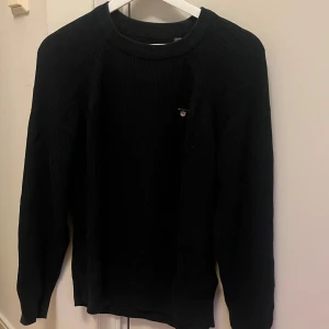 Mörk blå stickad tröja från Gant - Klassisk marinblå stickad tröja från Gant med rund halsringning och diskret logga på bröstet. Tröjan har ribbade muddar och lång ärm, perfekt för lager-på-lager-stil under kyliga dagar.