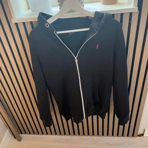Svart hoodie från Ralph Lauren - Svart hoodie från Ralph Lauren med dragkedja och huva. Klassisk design med vit dragkedja och rosa broderad logga på bröstet. Mjuk bomullskänsla och känguruficka framtill. Perfekt för chill dagar eller när du vill ha en clean streetstyle. M passar xs-s