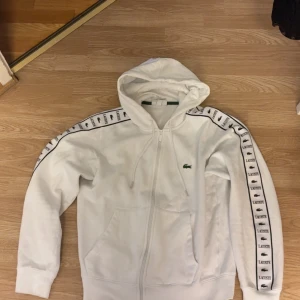 Vit hoodie från Lacoste med logoband - Snygg vit hoodie från Lacoste med dragkedja och huva. Ärmarna har breda logoband med Lacoste-tryck och krokodillogga längs hela sidan. Klassisk passform, kängurufickor och ribbade muddar. Perfekt för en sportig och clean look.