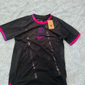 Svart PSG fotbollströja Nike - Snygg svart PSG fotbollströja från Nike med lila och rosa detaljer på krage och ärmslut. Tröjan har ett diskret blommönster i tyget och PSG-logga samt Nike swoosh i rosa framtill. Tillverkad i lätt och ventilerande material, perfekt för träning eller match.