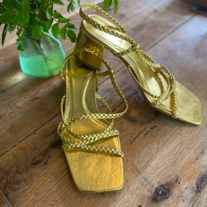 Guldiga sandaler med flätade remmar från Zara - Snygga sandaler i metallic guld från Zara med fyrkantig tå och flätade remmar. Skorna har en låg klack och öppen tå. Tillverkade i syntetmaterial med cool design och trendig vibe. Använd endast en gång till en specifik klänning. Sköna att bära dock