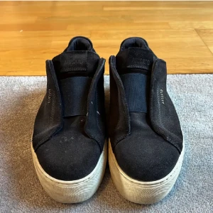 Svarta sneakers från Axel Arigato - Stilrena svarta sneakers från Axel Arigato med chunky vit platt sula. Ovandelen är i mocka och textil, utan synlig snörning för en clean look. Perfekta för dig som gillar minimalistisk stil och vill ha något som sticker ut. Skorna är sönder inuti!!!