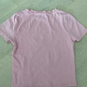Rosa t-shirt från Primark - Söt ljusrosa t-shirt från Primark med grönt tryck 'Hamptons Sports Club. Klassisk passform med korta ärmar och rund hals. Perfekt för en sportig och chill look.