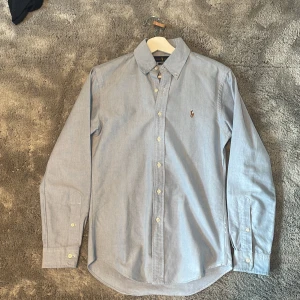 Ljusblå skjorta från Ralph Lauren - Klassisk ljusblå skjorta från Ralph Lauren i slim fit-modell. Skjortan har långa ärmar, knappar framtill och button down-krage. Broderad logga på bröstet. Perfekt för en clean och stilren look.