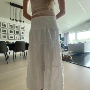 Vit långkjol från lager 157 - Säljer en vit långkjol med bred smock i midjan och volangiga lager. Kjolen är luftig och perfekt för varma dagar, med en enkel och stilren look. 