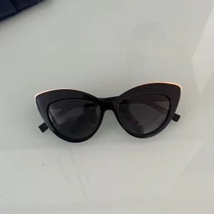 Svarta cat eye-solglasögon Le Specs - Snygga svarta cat eye-solglasögon från Le Specs med markerad guld båge och mörka glas. Ramen har en cool, spetsig form och en tunn gulddetalj längs ovankanten som ger extra edge. Perfekta för att lyfta din stil i sommar. Fodral i marinblått tyg och gummi medföljer.