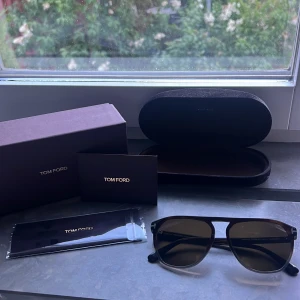 Tom Ford svarta solglasögon - Snygga solglasögon från Tom Ford med svarta bågar och mörka linser. Klassisk design med metall-detalj upptill och logga på skalmen. Modellen heter Jasper 02. Nypris ca 2600kr. Kommer med originalfodral, putsduk och box. Perfekta för att lyfta din stil i sommar. PRIS EJ HUGGET I STEN, kom gärna med bud. 