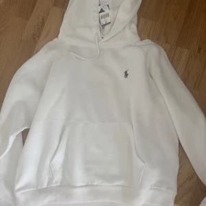 Vit hoodie från Polo Ralph Lauren XL - Vit hoodie från Polo Ralph Lauren i storlek XL. Klassisk design med broderad logga på bröstet, huva med snörning och stor magficka. Mjuk bomullskvalitet och ribbade muddar vid ärmslut och nederkant. Perfekt för en clean och stilren look. Prislappar kvar