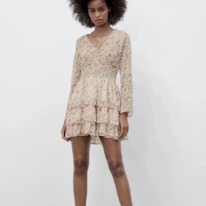 Blommig kortklänning med volang - Supersöt kortklänning med blommigt mönster i rosa och grönt på ljus botten. Klänningen har v-ringning, långa ärmar och smockad midja. Kjolen är volangig och ger en flowy känsla. Perfekt för varma dagar och somriga vibes!