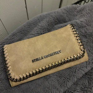 Beige plånbok Stella McCartney - Snygg beige plånbok (KP) Stella McCartney med coola kedjedetaljer runt kanterna. Plånboken har flera kortfack, fönster för ID och tryckknappsstängning. Perfekt för dig som vill ha en stilren och trendig accessoar i väskan.
