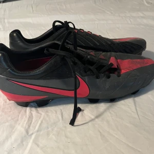 Nike T90 Laser fotbollsskor svart/röd - Nike T90 Laser fotbollsskor i svart och rött med coola grafiska detaljer och snörning. Skorna har dobbar för gräsplan och en sportig design med röd swoosh på sidan. Perfekta för dig som vill sticka ut på planen.  Skick enligt bilderna