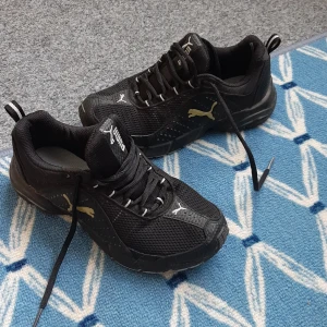 Svarta sneakers från Puma - Snygga svarta sneakers från Puma med guldigt logotyptryck på sidan. Ovandelen är i mesh och syntet för bra andningsförmåga. Skorna har snörning, platt sula och rund tå. Perfekta för dig som gillar sportig och stilren look.