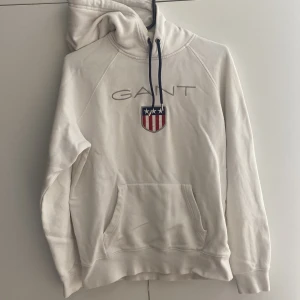 Vit hoodie från GANT med broderad logo - Vit hoodie från GANT med broderad logotyp och sköld på bröstet. Mörkblå snören i huvan och klassisk känguruficka framtill. Mjuk insida och ribbade muddar. Perfekt för en chill och stilren look.