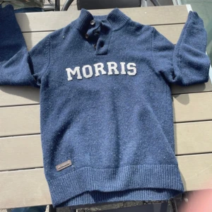 Blå ulltröja från Morris med knappar - Säljer en mörkblå stickad tröja från Morris med tydlig logga framtill. Tröjan har hög krage med knappar och ribbade muddar vid ärmslut och nederkant. Perfekt för kyliga dagar och snygg till jeans eller chinos.