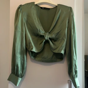 Zara blus/topp - Snygg grön omlottopp från Zara med lång ärm och knyt framtill. Toppen har croppad passform, djup v-ringning och breda manschetter. Materialet är glansigt och känns lyxigt, perfekt för att sticka ut.