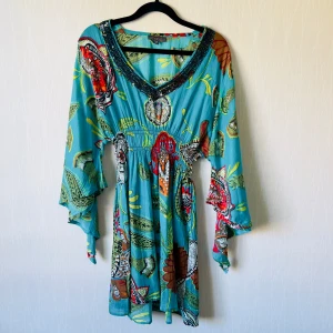 Blommig turkos klänning Tropical Melon - Färgstark kortklänning från Tropical Melon i turkos med stora blommiga mönster i rött, grönt och orange. V-ringad med paljettdetaljer vid halsen och vida, långa ärmar. Lätt och luftig i bomull, perfekt för sommarens varma dagar.