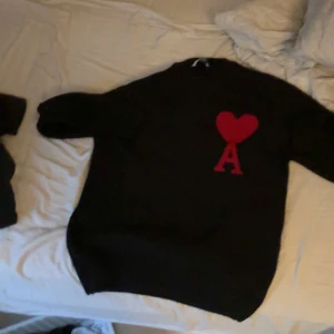 Svart stickad tröja Ami Paris hjärta - Svart stickad tröja från Ami Paris med ikoniskt rött hjärta och A på bröstet. Klassisk rund halsringning och lång ärm. Perfekt för dig som gillar stilrena plagg med en twist. Materialet är mjukt och håller värmen under kyliga dagar.