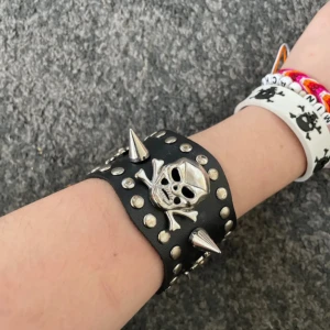 Svart nitarmband med dödskalle och spikes - Brett svart armband i skinnimitation med silverfärgade nitar, stora spikes och detaljer som dödskalle och kors. Perfekt för dig som gillar alternativ stil och vill sticka ut med accessoarer som har attityd.