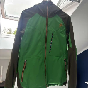  Elevenate skidjacka  - Snygg grön och mörkgrön vindjacka från Elevenate med huva och hel dragkedja framtill. Jackan har två fickor med dragkedja 