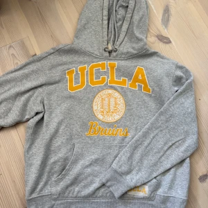 Grå UCLA hoodie med gul text - Snygg grå hoodie från UCLA med stor gul logga och texten Bruins på bröstet. Tröjan har huva med snörning och en stor magficka. Perfekt för dig som gillar college-stil och vill ha något bekvämt och stilrent.