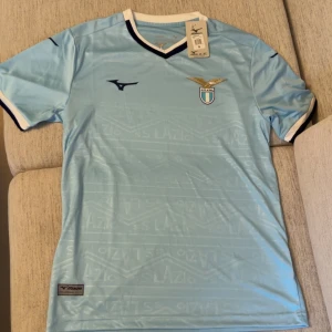 Ljusblå Lazio fotbollströja  - Snygg ljusblå Lazio fotbollströja från med klubbmärke på bröstet och mörkblå detaljer vid krage och ärmslut. Tröjan har subtila mönster med texten 'SS Lazio' i tyget och är tillverkad i ett lätt och ventilerande material. Perfekt för match eller träning.