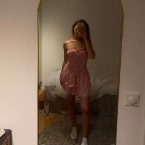 Rosa axelbandslös volangklänning - Supersöt rosa axelbandslös kortklänning med småblommigt mönster och volanger i kjolen samt inbyggda shorts under. Smockad överdel som sitter tajt och ger en snygg siluett.  Säljer endast då den tyvär inte kommer till använding! Den är sååååå fin och skön! Perfekt till sommaren 💖 St xs/S