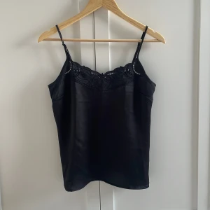Svart silkeslinne med spets från H&M - Supersnygg svart silkestopp från H&M med tunna axelband och dekorativ spets upptill. Sidenliknande material ger en lyxig känsla och toppen har en rak, enkel passform. Perfekt att styla med jeans eller kjol för en trendig look. Aldrig använd.