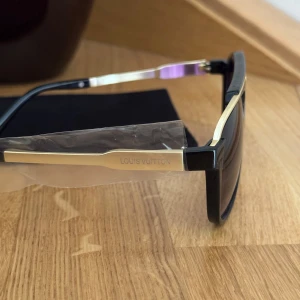 Louis Vuitton svarta solglasögon guld - Snygga solglasögon från Louis Vuitton med svart båge och guldfärgade detaljer. Glasen är mörka med en lätt toning och har en cool, sportig form. Märkets logga syns på skalmen och ovansidan har diskret LV-mönster. Perfekt accessoar för en trendig look.