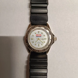 Prince quartzklocka med svart armband - Klassisk Prince quartzklocka med rund vit urtavla, tydliga siffror och gulddetaljer. Boetten är i silverfärgad metall och armbandet är svart och ribbat i plast. Vattentät upp till 3 ATM och har en sportig look med markerad bezel.