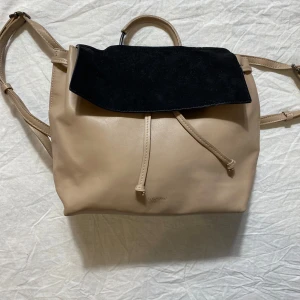 Beige och svart ryggsäck från Don Donna - Snygg ryggsäck i beige skinnimitation från Don Donna med svart mockadetalj på locket. Väskan har justerbara axelremmar, handtag upptill och stängs med snörning och magnetknapp. Perfekt för dig som vill ha en stilren och praktisk väska.