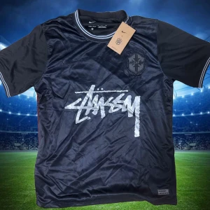 Svart Brasil x Stüssy fotbollströja Nike - Snygg svart fotbollströja från Nike i samarbete med Stüssy och Brasil. Tröjan har korta ärmar, ribbade muddar med vita detaljer och stor Stüssy-logga framtill. Broderat Brasil-märke på bröstet och fem stjärnor i nacken. Tillverkad i lätt och ventilerande material.storlek S,M