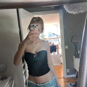 Svart bandeau tubtopp med pärldetalj - Säljer en svart bandeau tubtopp med snygg rynkad effekt och en liten pärldetalj framtill vid uringningen. Toppen är axelbandslös och sitter tight, perfekt till jeans eller kjol för en trendig look.