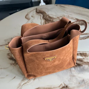 Brun mocka axelväska från Prada - Säljer en snygg brun axelväska från Prada i lyxig mocka med guldiga metalldetaljer och klassisk logga framtill. Väskan har flera fack och en justerbar axelrem, perfekt för dig som vill ha både stil och funktion. Rymmer allt du behöver till vardagen.