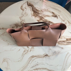 Brun flätad handväska från bottega veneta - Snygg brun handväska i flätad design med rejäla handtag. Väskan är tillverkad i skinn och har ett stort fack med innerficka som stängs med dragkedja och magnetknapp. Perfekt för dig som vill ha en stilren och rymlig väska till vardagen.