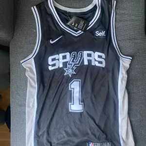 San Antonio Spurs basketlinne  - Denna vara är en replika. Helt oanvänt och bara provad med tagg även. Säljer eftersom den är för stor på mig. Står att varan är L men passar mer som en XL med skum passform. Victor Wembanyama tröja. Är 181 men den var för lång för mig