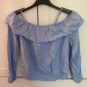 Ljusblå offshoulder blus  - Supersöt ljusblå blus med offshoulder och volang runt hela ringningen. Långa ärmar och smock i nederkant och ärmslut. 