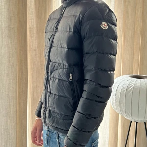 Svart Moncler Acorus jacka herr - Snygg svart dunjacka från Moncler med klassisk quiltad design och diskret logga på ärmen. Jackan har dragkedja framtill, två fickor med dragkedja och hög krage. Perfekt för kyliga dagar och ger en clean, stilren look.