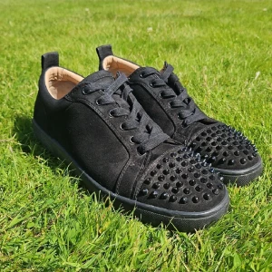 Christian Louboutin svarta sneakers med nitar - Svarta sneakers i mocka från Christian Louboutin med ikoniska röda sulor och svarta nitar på tån. Skorna har snörning och rund tå, samt lyxig känsla med läderdetaljer invändigt. Perfekta för dig som vill sticka ut med exklusiv streetstyle.