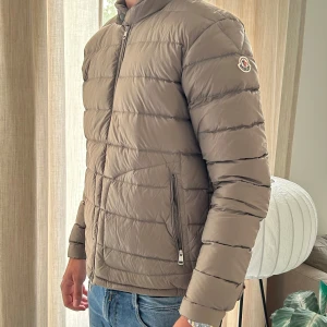 Beige Moncler Acorus jacka herr - Snygg beige dunjacka från Moncler med klassisk logga på ärmen. Jackan har quiltad design, dragkedja framtill och två fickor med dragkedja. Perfekt för kyliga dagar och ger en clean, stilren look. Passar dig som gillar exklusiva märken och streetstyle.