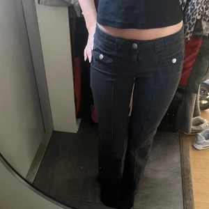 Svarta lågmidjade bootcut jeans - Säljer ett par svarta jeans med låg midja och bootcut-ben. Byxorna har snygga fickdetaljer framtill med silverfärgade knappar och markerade sömmar längs benen. Perfekta för dig som gillar en retro vibe och vill ha ett par jeans som sticker ut. Jag är 164cm