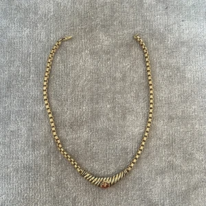 Guldfärgat halsband med kedja - Snyggt halsband i guldfärg med fyrkantig kedja och ett dekorativt, böjt hänge framtill. Hänget har räfflade detaljer och en liten infattad sten i mitten. Har fått halsbandet i present och vet därför inte märket, säljer det då det inte är min stil💕