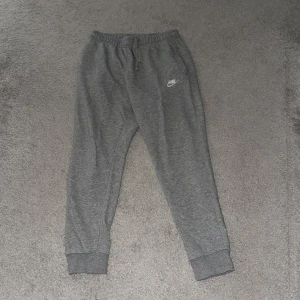 Grå mjukisbyxor från Nike - Gråa mjukisbyxor från Nike med klassisk logga på vänster ben. Byxorna har resår och snörning i midjan samt muddar vid bensluten. Perfekta för chill eller träning, tillverkade i mjukt bomullsmaterial.