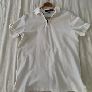 Vit pikétröja från Polo Ralph Lauren - Klassisk vit pikétröja från Polo Ralph Lauren med korta ärmar, krage och half zip-dragkedja framtill. Tröjan har broderad logga på bröstet och är gjord i bomull för en skön och stilren look. Nästan oanvänd. Nypris 1499