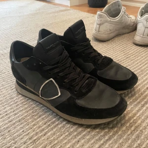 Svarta sneakers från Philippe Model - Snygga svarta sneakers från Philippe Model med klassisk design. Skorna har ovandel i skinn och mocka, svarta snören och en kontrasterande vit detalj på sidan. Perfekta för dig som gillar stilrena och sportiga skor.