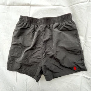 Svarta badshorts från Polo Ralph Lauren - Svarta badshorts från Polo Ralph Lauren med elastisk midja och röd broderad logga på benet. Shortsen har innerbyxa i mesh och en bakficka med lock. Perfekta för stranden eller poolen, klassisk och stilren design.
