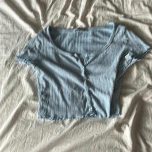Ljusblå ribbad topp från FB Sister - Supersöt ljusblå croppad topp från FB Sister med ribbat tyg och små knappar framtill. Toppen har korta ärmar och en lätt v-ringad halsringning. Perfekt för dig som gillar en chill och trendig stil.