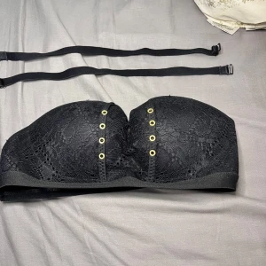 Självhäftande spets behå bra push up  - Säljer en svart bandeau tubtopp med snygg spets och guldfärgade öljetter framtill. Toppen är axelbandslös men levereras med avtagbara svarta axelband. Insidan har vaddering och silikon för extra stöd. Perfekt till outfits där du vill visa axlarna.