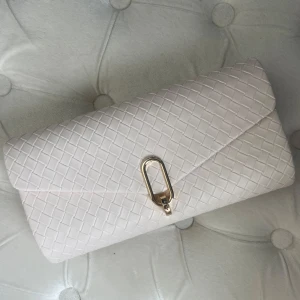 Vit kuvertväska / clutch med kedja - Snygg cremevit kuvertväska med flätad struktur och guldfärgad kedja, vilket går att ta bort och göra till clutch. Väskan har ett elegant guldspänne framtill. Perfekt till bal. Helt oanvänd.