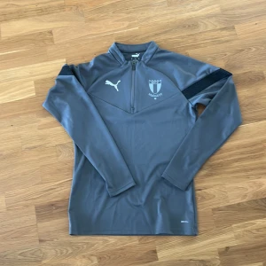 Grå Malmö FF träningströja Puma S - Snygg grå långärmad träningströja från Puma med Malmö FF-logga på bröstet. Tröjan har halv dragkedja framtill och svarta detaljer på axlarna. Tillverkad i funktionsmaterial som andas, perfekt för träning eller match.